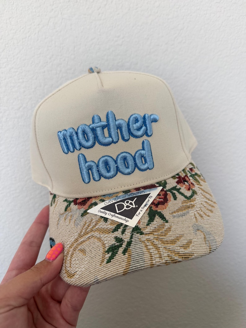 Puede incluir: Gorra de b&eacute;isbol beige con la palabra "motherhood" bordada en azul. La visera presenta un dise&ntilde;o de tapiz floral y una etiqueta "David & Young".