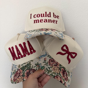 Puede incluir: Tres gorras de b&eacute;isbol color crema con viseras estampadas con flores. Una gorra tiene el texto "I could be meaner" bordado en borgo&ntilde;a. Otra dice "MAMA" en borgo&ntilde;a. La tercera gorra presenta un lazo borgo&ntilde;a.