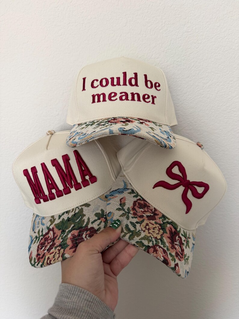 Puede incluir: Tres gorras de b&eacute;isbol color crema con viseras estampadas con flores. Una gorra tiene el texto "I could be meaner" bordado en borgo&ntilde;a. Otra dice "MAMA" en borgo&ntilde;a. La tercera gorra presenta un lazo borgo&ntilde;a.