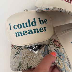 May include: Off-white baseball cap with the text "I could be meaner" embroidered in teal. The brim features a floral tapestry design. A second cap with the text "I could mean" is partially visible.