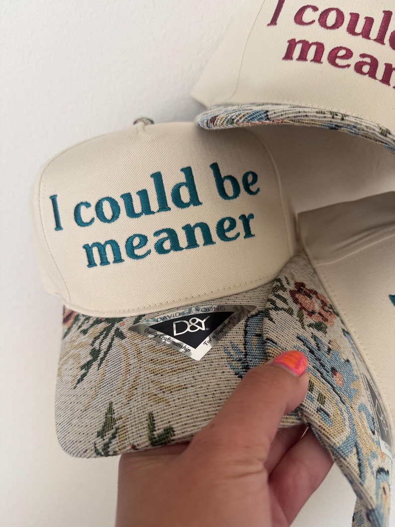 May include: Off-white baseball cap with the text "I could be meaner" embroidered in teal. The brim features a floral tapestry design. A second cap with the text "I could mean" is partially visible.