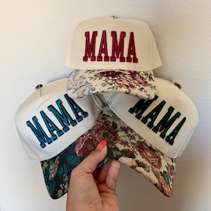 Puede incluir: Tres gorras de b&eacute;isbol color crema con la palabra "MAMA" bordada en rojo, verde azulado y verde. Las viseras presentan estampados florales en varios colores. Las gorras se muestran sobre un fondo blanco.