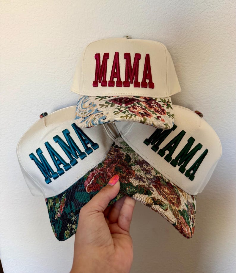 Puede incluir: Tres gorras de b&eacute;isbol color crema con la palabra "MAMA" bordada en rojo, verde azulado y verde. Las viseras presentan estampados florales en varios colores. Las gorras se muestran sobre un fondo blanco.
