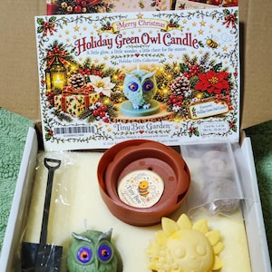 Puede incluir: Una caja de regalo que contiene una vela verde con forma de búho, una vela amarilla con forma de girasol, una maceta pequeña color terracota con una pegatina de Tiny Bee Garden, una pala de plástico negra y una bolsa con dos objetos redondos.