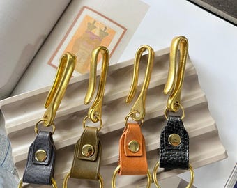 May include: Four keychains featuring gold-toned metal hooks and rings attached to leather tabs. Each tab is secured with a gold-toned metal snap button. The leather tabs are in shades of dark gray, olive green, orange, and textured black.