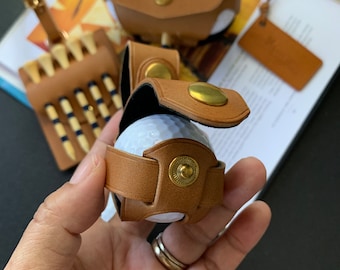 May include: A collection of golf accessories in shades of brown leather. The image features a golf ball holder, a tee holder, and a small pouch, all with brass hardware. The golf ball holder is secured around a white golf ball.
