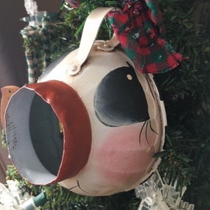 May include: A white ceramic ornament shaped like a snowman's head with a red, green, and white plaid fabric scarf. The ornament has black painted eyes, a red painted nose, and a pink painted cheek.