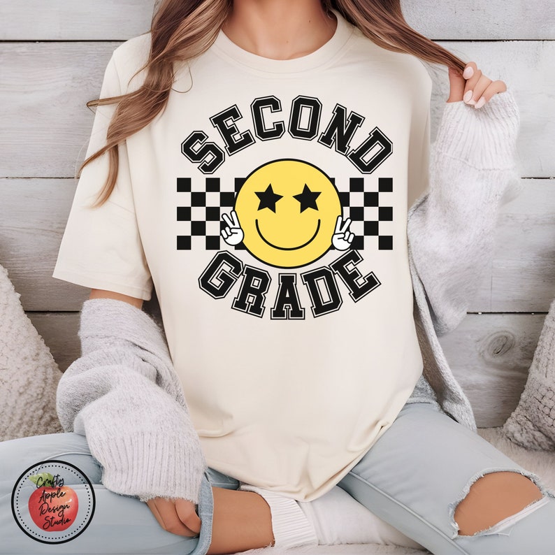 Puede incluir: Una camiseta blanca con el texto "SECOND GRADE" en negro con una cara sonriente amarilla con ojos de estrella negra y signos de paz. La cara sonriente est&aacute; sobre un fondo a cuadros blanco y negro.
