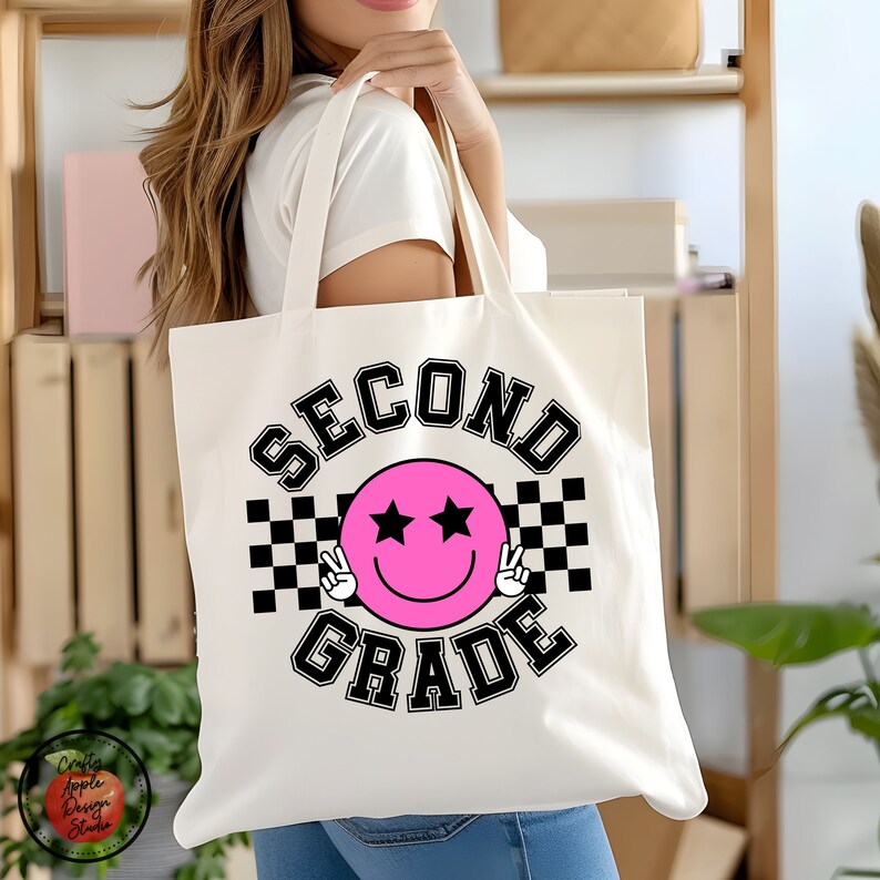 Puede incluir: Bolsa de tela blanca con un dise&ntilde;o a cuadros blanco y negro y una cara sonriente rosa con estrellas por ojos. El texto "SECOND GRADE" est&aacute; en un dise&ntilde;o arqueado blanco y negro sobre la cara sonriente.