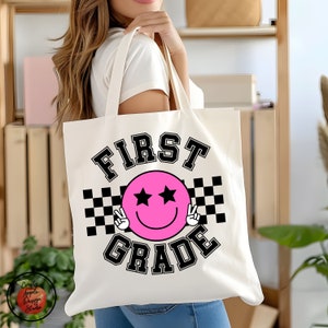 Może przedstawiać: Biała torba z pł&oacute;tna z czarnym tekstem, kt&oacute;ry brzmi "FIRST GRADE" w stylu uniwersyteckim. R&oacute;żowa buźka z czarnymi gwiazdkami w oczach i znakami pokoju w dłoniach znajduje się w środku torby. Czarno-biały wz&oacute;r w kratkę znajduje się za buźką.