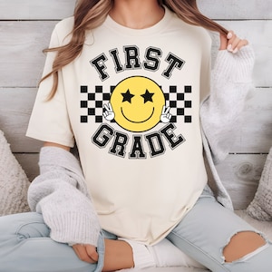 Może przedstawiać: Biała koszulka z czarno-białym wzorem w kratkę i ż&oacute;łtą buźką z czarnymi gwiazdkami i znakami pokoju. Tekst "FIRST GRADE" jest napisany czarnym kolorem nad i pod buźką.