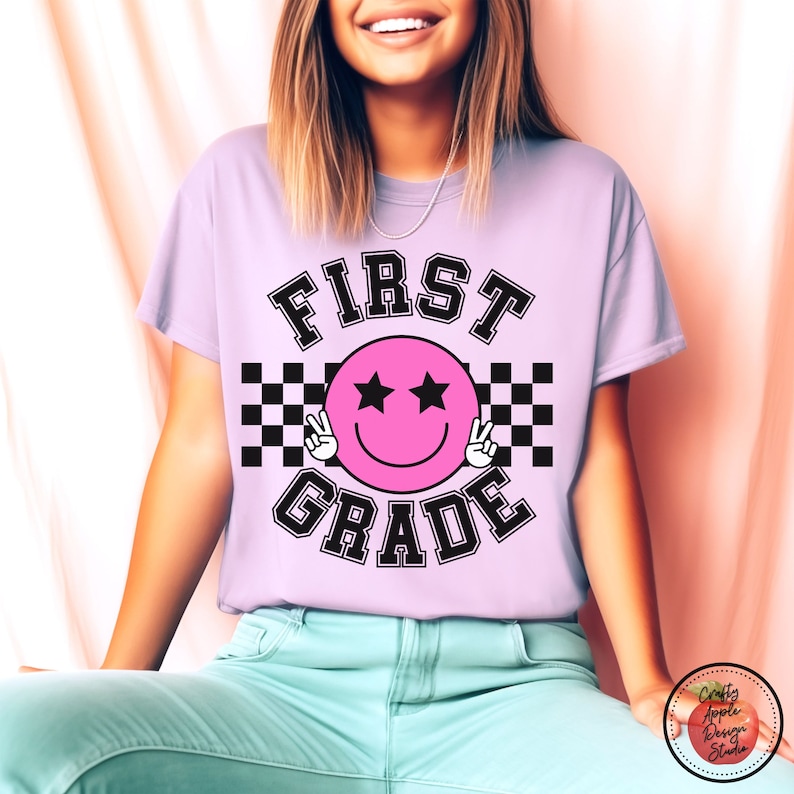 Może przedstawiać: Jasno-fioletowy t-shirt z napisem "FIRST GRADE" w czarnych, obwiedzionych literach. W środku napisu znajduje się r&oacute;żowa buźka z czarnymi gwiazdkami w oczach i znakami pokoju. Za buźką znajduje się czarno-biały wz&oacute;r w kratkę.