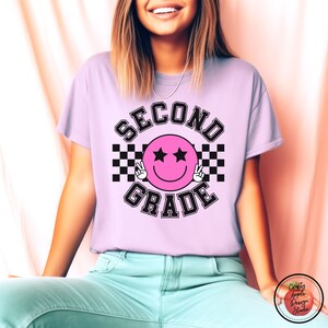 Puede incluir: Una camiseta de color morado claro con un dise&ntilde;o a cuadros en blanco y negro y el texto "SECOND GRADE" en una fuente con contorno negro. Una cara sonriente rosa con ojos negros y signos de paz se encuentra en el centro del dise&ntilde;o.