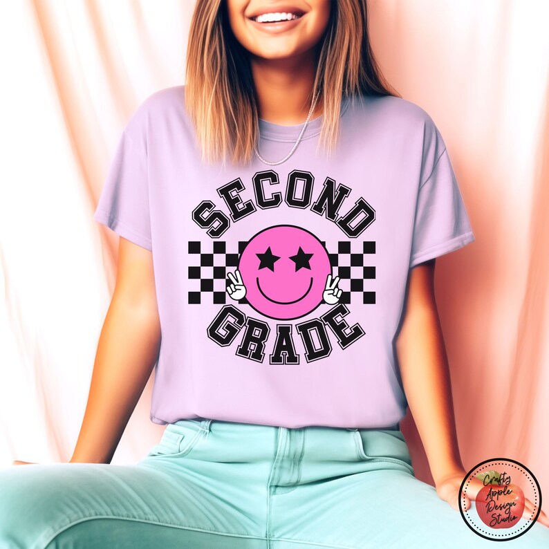 Puede incluir: Una camiseta de color morado claro con un dise&ntilde;o a cuadros en blanco y negro y el texto "SECOND GRADE" en una fuente con contorno negro. Una cara sonriente rosa con ojos negros y signos de paz se encuentra en el centro del dise&ntilde;o.
