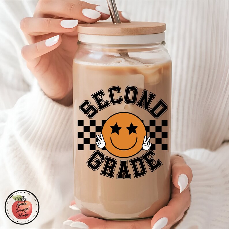 Puede incluir: Un vaso de vidrio transparente con tapa de madera y un dise&ntilde;o a cuadros en blanco y negro con el texto "SECOND GRADE" y una carita sonriente con dos estrellas. El vaso est&aacute; lleno de caf&eacute; helado.