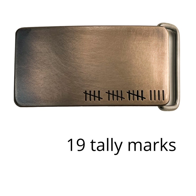 May include: A rectangular metal belt buckle with a brushed bronze finish. The buckle features nineteen tally marks etched on the surface. The text "19 tally marks" is below the buckle.
