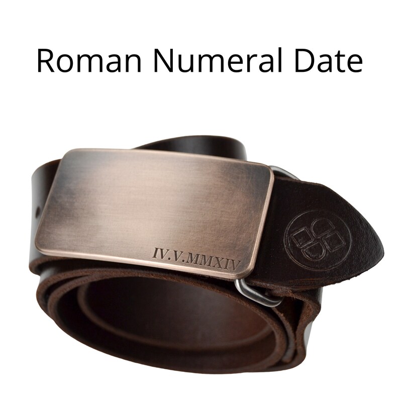 May include: A dark brown leather belt with a rectangular bronze-colored buckle. The buckle has the Roman numeral date IV.V.MMXIV engraved on it. The belt is rolled up, showing the texture of the leather.
