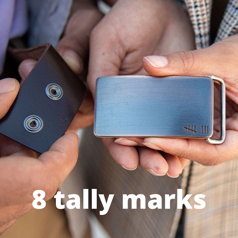 May include: A rectangular metal belt buckle with a brushed finish and a silver-colored frame. The buckle has eight tally marks etched on the lower right corner. The image also shows a brown leather belt strap with two metal snap fasteners. The text "8 tally marks" is at the bottom.