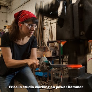 Op de afbeelding: Een vrouw met veiligheidsbril en een rode bandana bedient een grote metalen kracht hamer in een werkplaats.