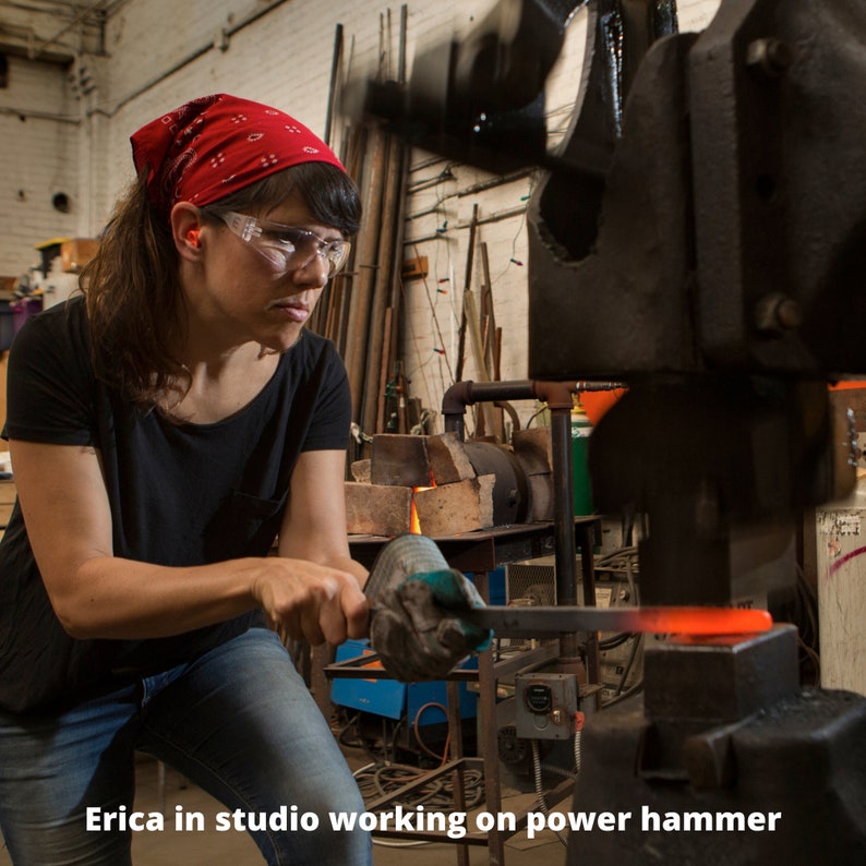 Op de afbeelding: Een vrouw met veiligheidsbril en een rode bandana bedient een grote metalen kracht hamer in een werkplaats.