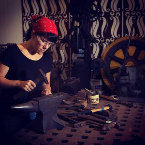 May include: A person wearing a red bandana and safety glasses working in a workshop. They are using a hammer and anvil. Various tools are scattered on a metal surface. The background features industrial equipment and a patterned wall.