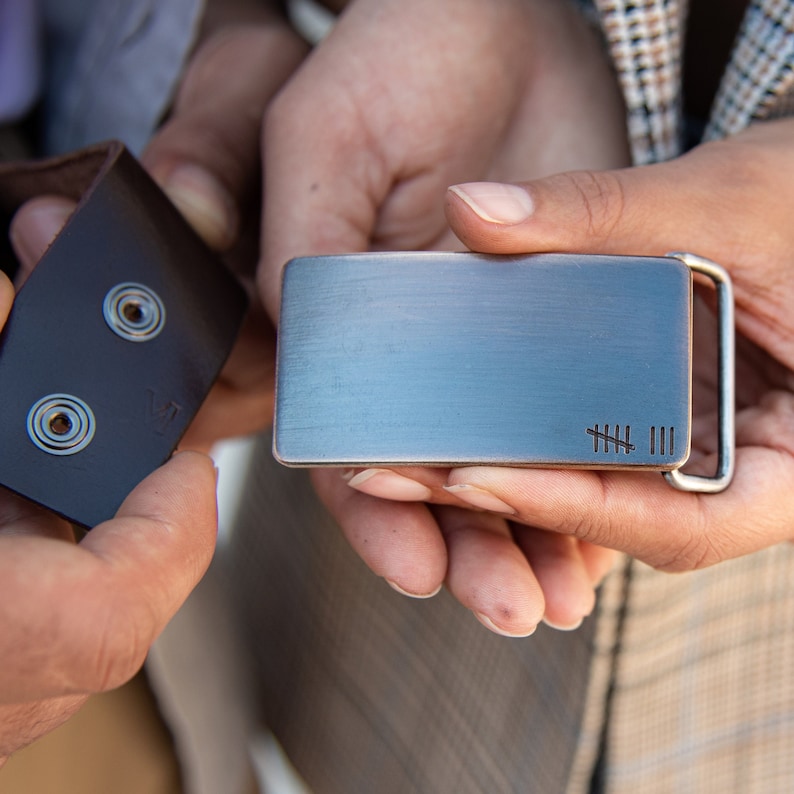 A bronze metal belt buckle with a brushed finish and a small engraved design of 8 tally marks to represent 8 years of marriage. The buckle is rectangular in shape and a snapped belt is shown.