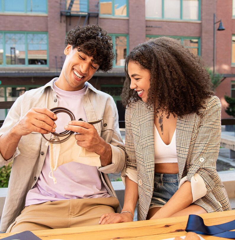 Smiling couple looking at iron anniversary belt buckle he has just been gifted in an outdoor, coffee shop city setting.