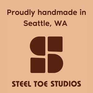 May include: A logo design on a peach background. The text reads "Proudly handmade in Seattle, WA" in a dark brown font. Below the text is a dark brown graphic of four connected shapes. The words "STEEL TOE STUDIOS" are at the bottom in a dark brown font.