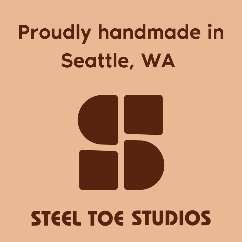 May include: A logo design on a peach background. The text reads "Proudly handmade in Seattle, WA" in a dark brown font. Below the text is a dark brown graphic of four connected shapes. The words "STEEL TOE STUDIOS" are at the bottom in a dark brown font.