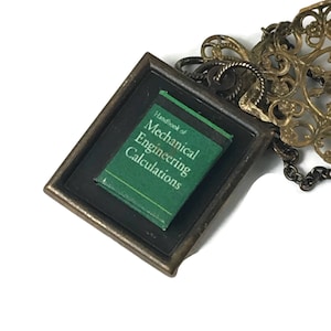 May include: A bronze-toned pendant featuring a miniature book with the title "Handbook of Mechanical Engineering Calculations" on a green cover. The pendant has a rectangular frame and an ornate chain.