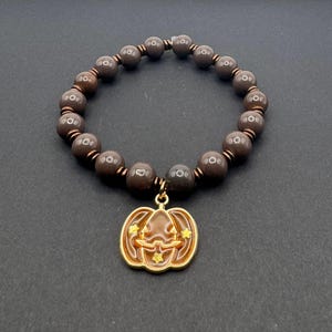Puede incluir: Un brazalete de cuentas marrones con un dije de calabaza dorado. La calabaza tiene una cara tallada y estrellas doradas.