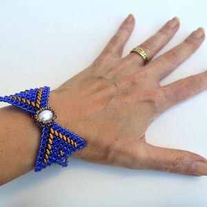 Könnte beinhalten: Ein blaues Perlenarmband mit goldenen Akzenten und einer zentralen perlenartigen Perle. Das Armband hat die Form einer Schleife. Das Armband befindet sich an einem Handgelenk.