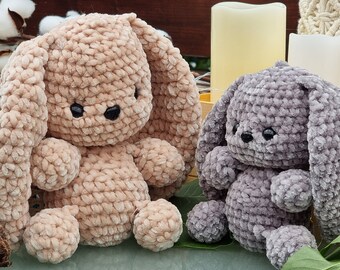 May include: Two crocheted bunny plushies, one in a light brown color and the other in a dark gray color. Both plushies have long ears and black eyes.