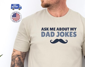 May include: A light beige t-shirt with the text "Ask me about my dad jokes" in blue lettering. A blue mustache graphic is below the text.