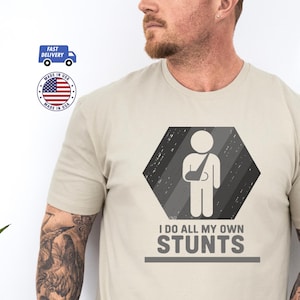 May include: A light beige t-shirt with a graphic of a person with an arm in a sling inside a gray hexagon. The text "I DO ALL MY OWN STUNTS" is below the graphic. The shirt also has "Made in USA" and "Fast Delivery" logos.