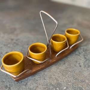 May include: A set of four small, yellow ceramic cups with a wooden base and a metal stand. The cups are arranged in a row on the base, and the stand is designed to hold the cups upright.