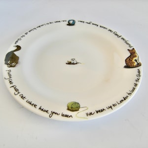 May include: White ceramic plate with a whimsical design featuring a cat, mouse, and yarn ball. The plate has the text "Pussy cat pussy cat where have you been, I've been up to London to look at the Queen" around the rim.