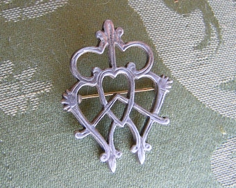 May include: A silver-toned brooch with an ornate design featuring three interconnected hearts and a crown-like element at the top. The brooch has a pin closure on the back and is likely made of metal.