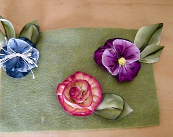 May include: Three silk ribbon flowers on a green felt background. The flowers are a blue and white flower, a purple and white flower, and a pink and white flower.