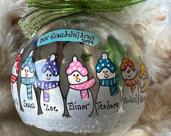 May include: A clear glass Christmas ornament with a green ribbon. The ornament is decorated with painted snowmen wearing colorful hats and scarves. The names of the grandchildren are written below each snowman. The text "our grandchildren 2024" is written on the top.