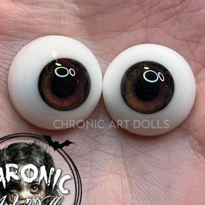 Puede incluir: Dos ojos de muñeca con iris marrones y esclerótica blanca. Los ojos tienen un acabado brillante y están sobre un fondo claro. El texto "CHRONIC ART DOLLS" es visible.