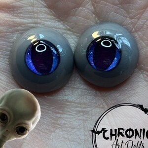May include: Two gray and blue doll eyes with a purple iris and a black pupil. The eyes are shiny and have a glitter effect.