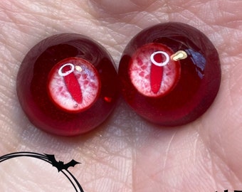 May include: Two red glass eyes with white pupils and a gold detail. The eyes are on a hand.