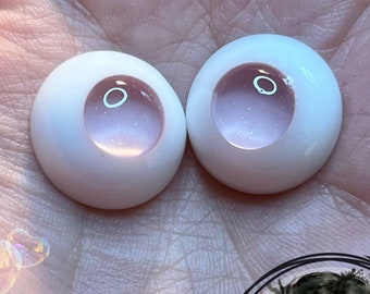 Peut inclure: Deux yeux de poupée blancs avec des iris rose et argent pailletés. Les yeux sont sur une surface blanche.