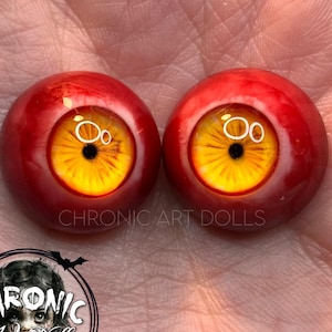 May include: Two doll eyes with a vibrant design. The irises are yellow with black pupils, set within a red sclera. The text "CHRONIC ART DOLLS" is visible. The eyes are round and appear to be made of glass or resin.
