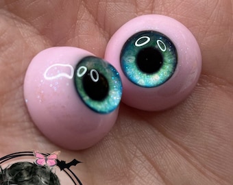 Peut inclure: Deux grands yeux de poupée ronds, bleus et verts avec des pupilles noires. Les yeux sont sur un fond rose.