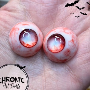 Może przedstawiać: Dwa realistycznie wyglądające oczy dla lalki z czerwono-białym marmurem. Oczy są szklane i mają realistyczną tęcz&oacute;wkę i źrenicę.