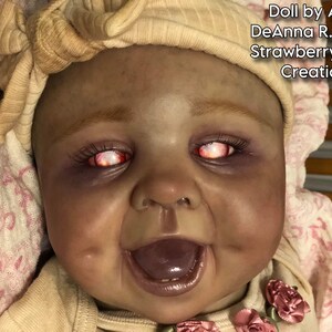 Może przedstawiać: Zbliżenie na reborn doll o ciemnej sk&oacute;rze, r&oacute;żowo-białej opasce na głowę i jaskrawoczerwonych oczach. Lalka uśmiecha się z otwartymi ustami, ukazując zęby i fioletowy język. Lalka jest ubrana w biało-r&oacute;żowy str&oacute;j z kwiatowym wzorem.