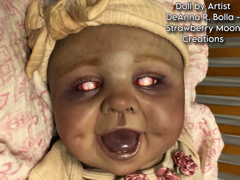 Może przedstawiać: Zbliżenie na reborn doll o ciemnej sk&oacute;rze, r&oacute;żowo-białej opasce na głowę i jaskrawoczerwonych oczach. Lalka uśmiecha się z otwartymi ustami, ukazując zęby i fioletowy język. Lalka jest ubrana w biało-r&oacute;żowy str&oacute;j z kwiatowym wzorem.
