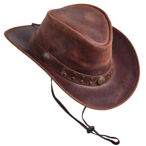 May include: A brown leather cowboy hat with a distressed finish. The hat features a wide brim, a decorative band with rivets, and a chin strap. The hat is a classic Western style.